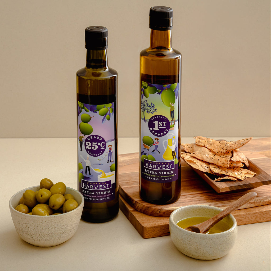 Two bottles of Harvest Olive Virgin Oil stand on a wooden board with olive green olives in a bowl, crackers on a small tray, and a bowl of olive oil with a spoon, set against a warm beige background.
