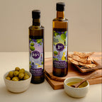Two bottles of Harvest Olive Virgin Oil stand on a wooden board with olive green olives in a bowl, crackers on a small tray, and a bowl of olive oil with a spoon, set against a warm beige background.