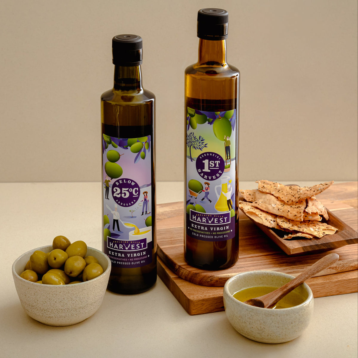 Two bottles of Harvest Olive Virgin Oil stand on a wooden board with olive green olives in a bowl, crackers on a small tray, and a bowl of olive oil with a spoon, set against a warm beige background.
