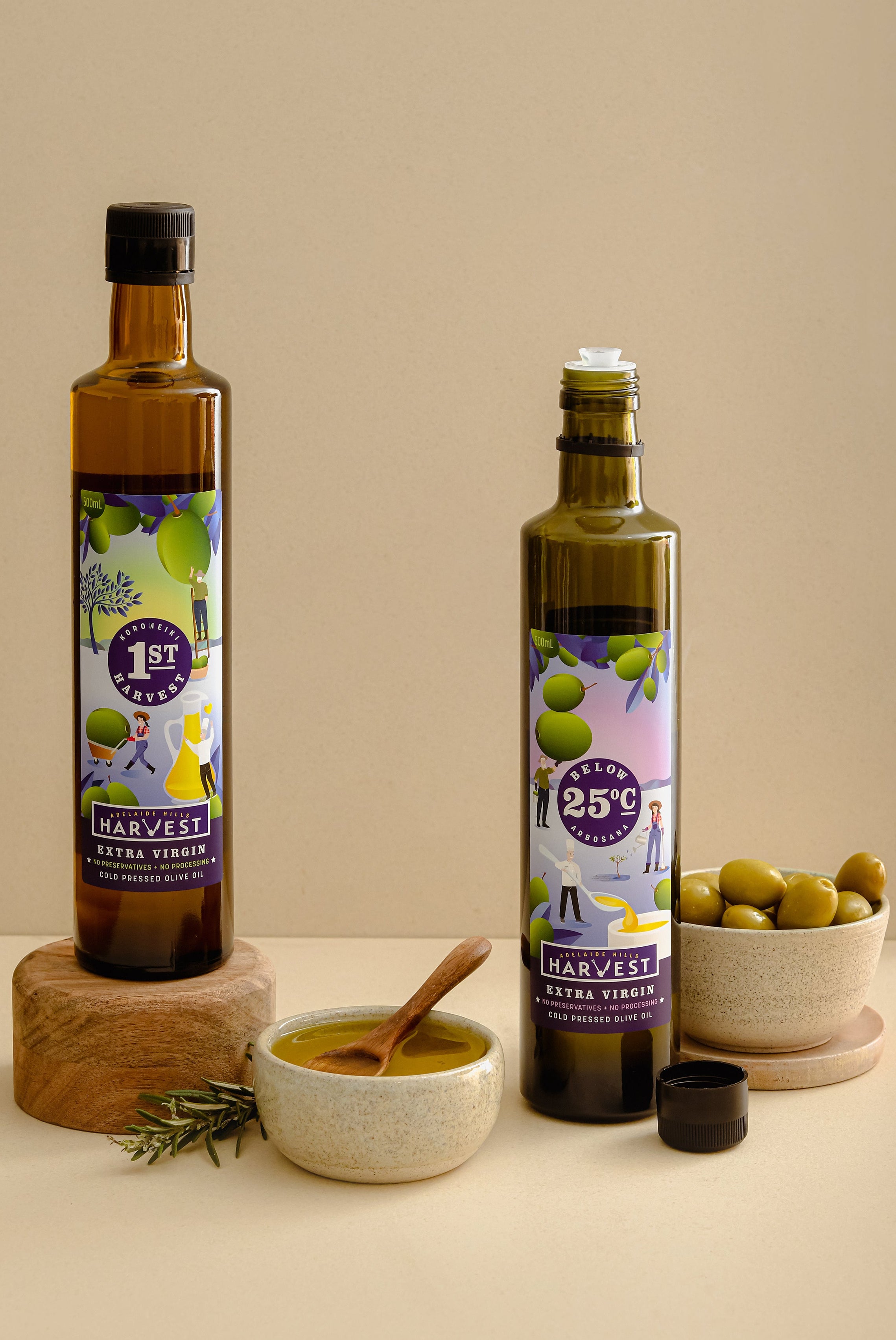 Two glass bottles of Harvest 1st Press extra virgin olive oil stand on a beige studio background, styled with bowls of olives, a spoonful of olive paste, and a small sprig of rosemary. One bottle is on a round wooden riser.