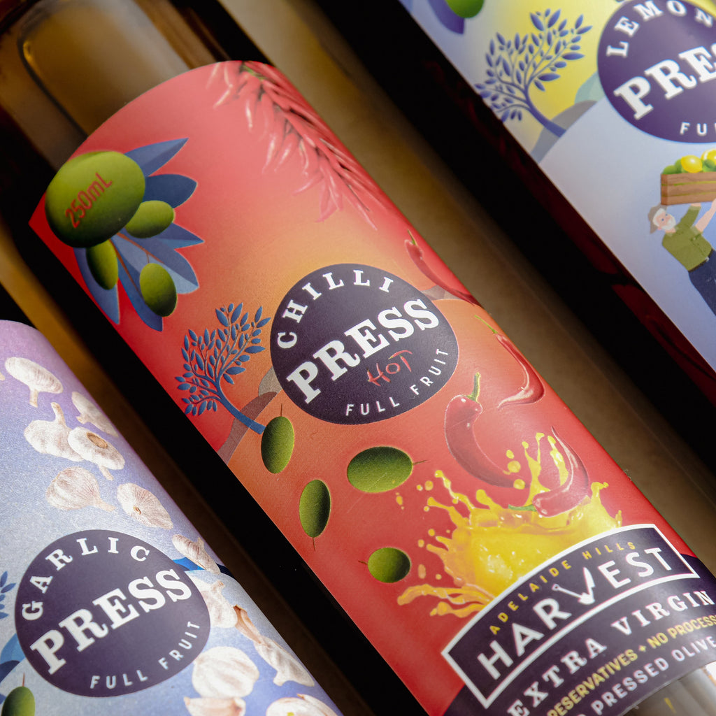 Close-up of olive oil bottles with colorful labels featuring brand names like 'Harvest' and 'Chilli Press'.