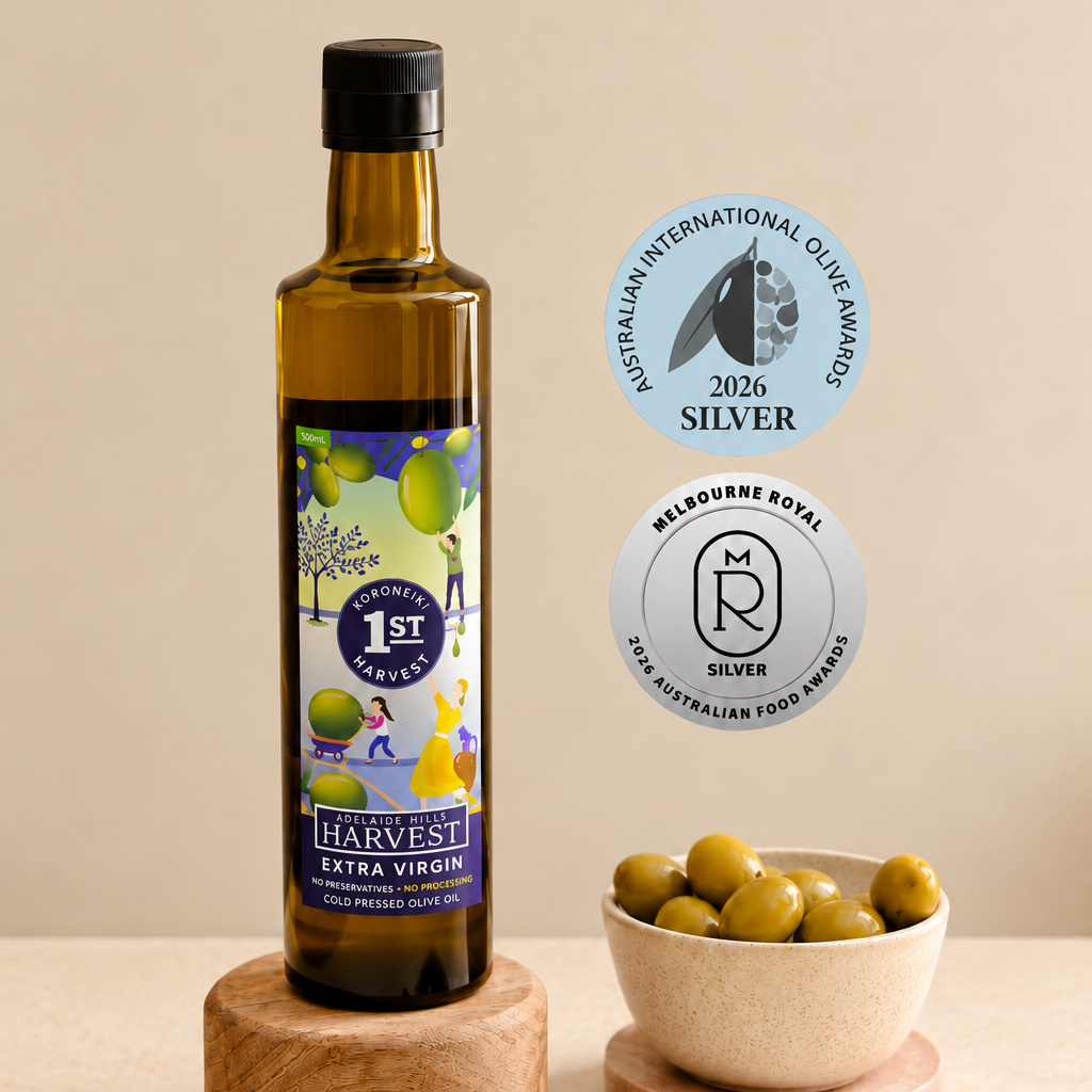 A bottle of Harvest Extra Virgin olive oil displayed on a wooden pedestal, with a bowl of whole olives, a small dish of oil, and rosemary sprigs on a beige surface. Two award badges are shown to the right: the Australian International Olive Awards 2023 Silver and the Melbourne Royal Gold Awards Silver.
